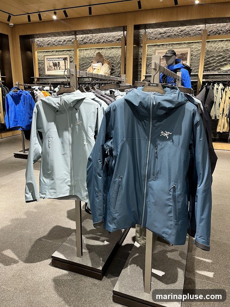 A retail display featuring several Arc'teryx jackets on hangers. Two jackets are prominently displayed in the foreground, one light blue and one dark blue. In the background, more jackets and apparel are visible on racks, along with mannequins dressed in outdoor gear. The store interior has wooden accents and track lighting.