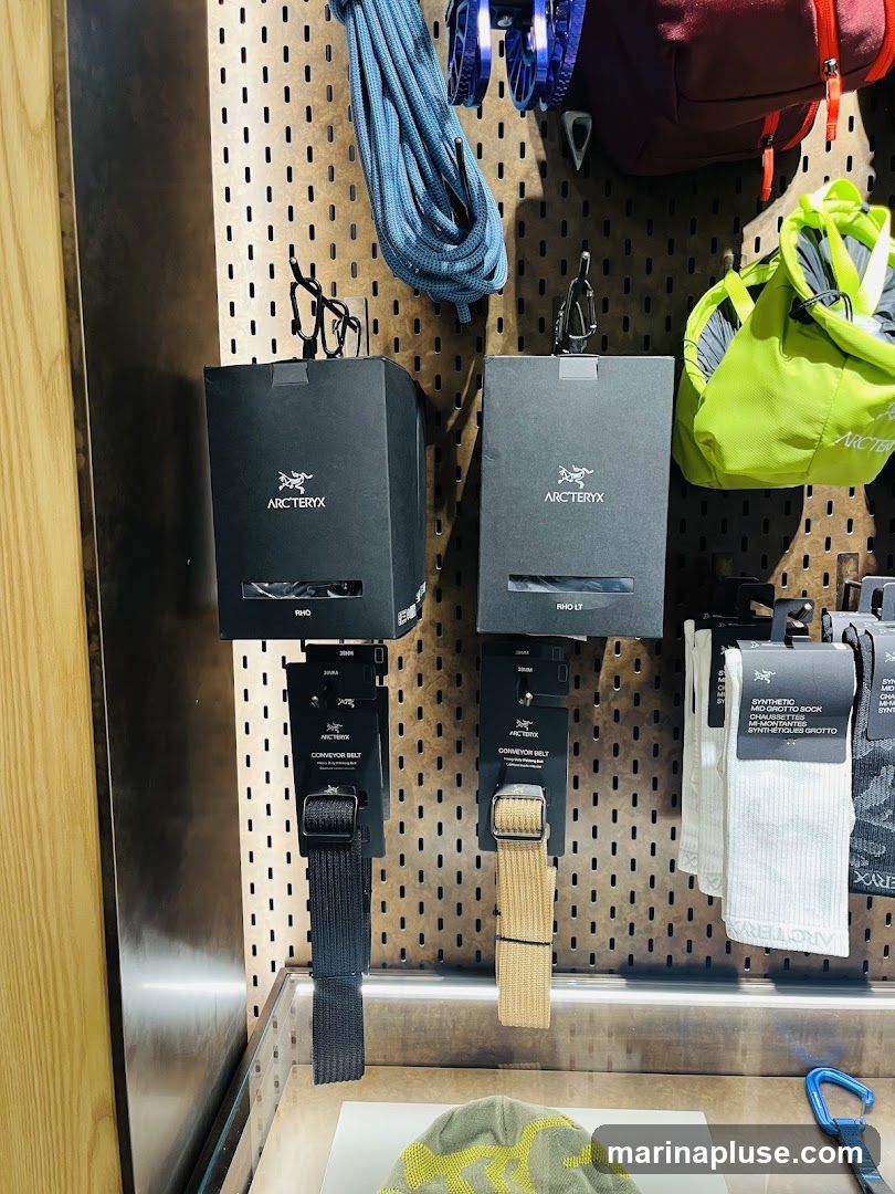 A retail display featuring Arc'teryx products, including two belts and several pairs of socks. The belts are black and tan, labeled as "CONVEYOR BELT Heavy Duty Webbing Belt". Above the belts are black boxes with the Arc'teryx logo. To the right, white and grey socks are displayed, with one pair labeled "SYNTHETIC MID GROTTO SOCK". Blue rope and backpacks are visible in the background.