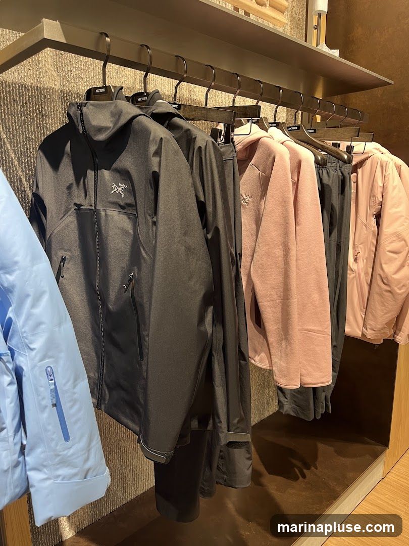 A rack of outdoor jackets and apparel is displayed on hangers. The jackets are in various colors including light blue, black, and pink. The black jackets feature the Arc'teryx logo. The apparel is neatly arranged on a shelf with a textured wall behind it.