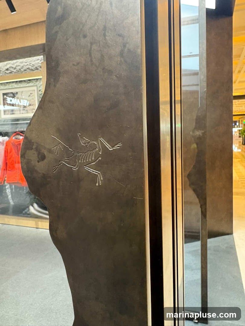 A close-up view of a dark, textured surface with an etched logo of a skeletonized human figure. The logo appears to be the Arc'teryx bird logo. In the background, through a glass window, racks of bright orange jackets are visible, suggesting an apparel store. The overall impression is of a modern, minimalist retail environment.