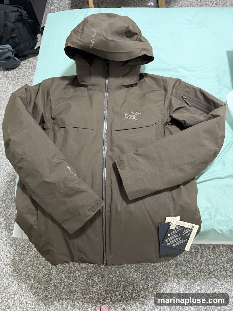 A brown Arcteryx Gore-Tex jacket is laid flat on a light blue surface. The jacket has a hood with "ARCTERYX" printed on it. The Arcteryx logo is visible on the chest. There are zippers and velcro closures on the jacket. Several tags are attached to the lower right side of the jacket.
