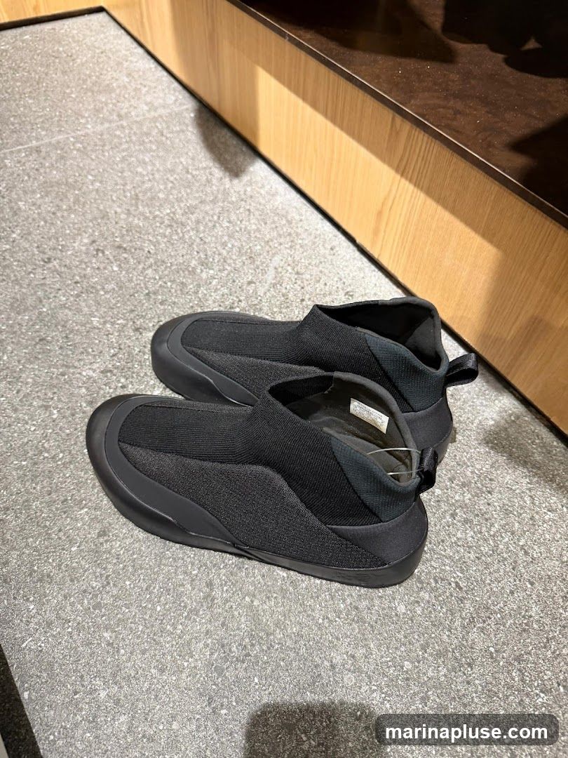 A pair of black slip-on shoes with a textured fabric upper and a smooth sole are displayed on a speckled gray floor. Behind the shoes, a wooden display shelf with a dark brown top is visible.