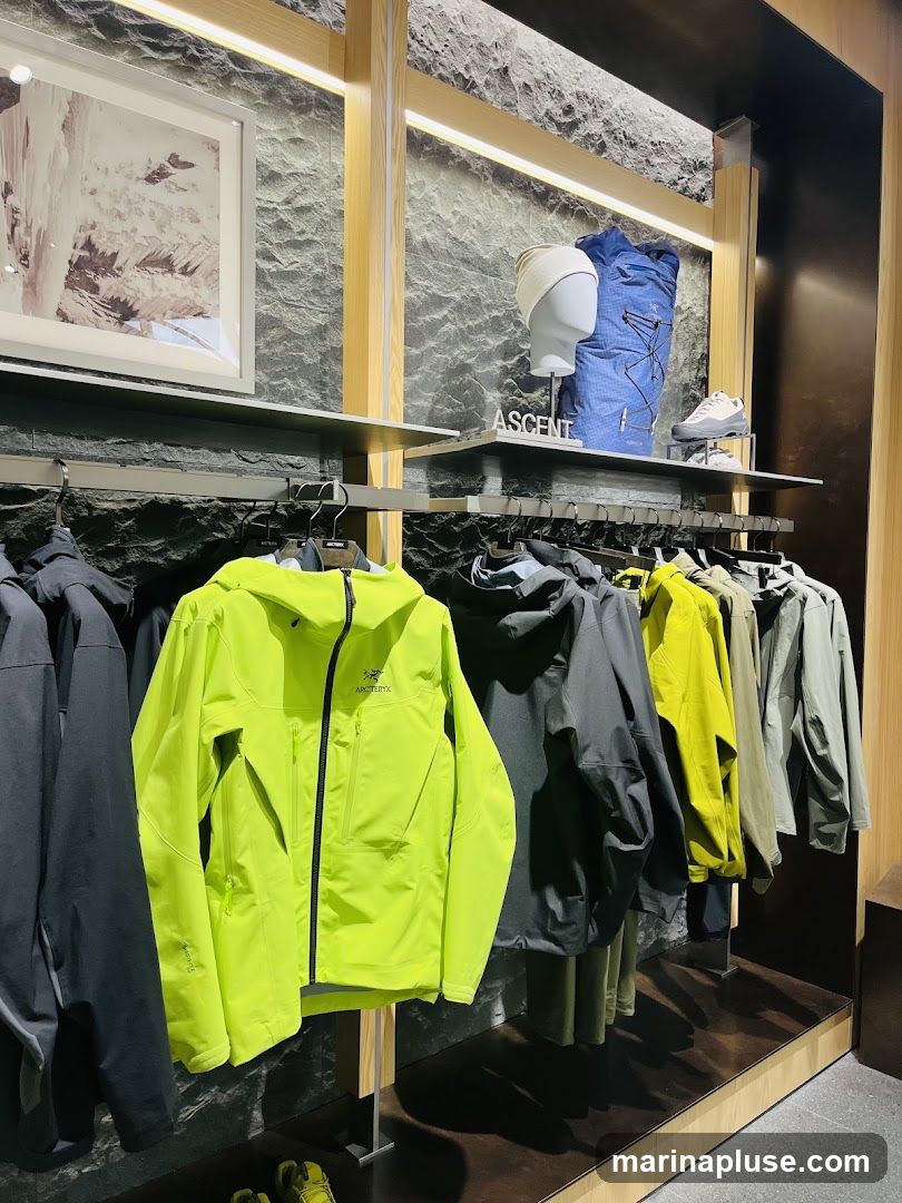 A retail display featuring various outdoor jackets and accessories. In the foreground, several jackets are hung on a rack, including a bright lime green jacket with the Arc'teryx logo. Behind the jackets, shelves display a mannequin head wearing a beanie, a blue backpack, and a pair of hiking shoes. A framed picture of a natural landscape hangs on the wall above the shelves. The word "ASCENT" is visible on a small sign on the shelf.