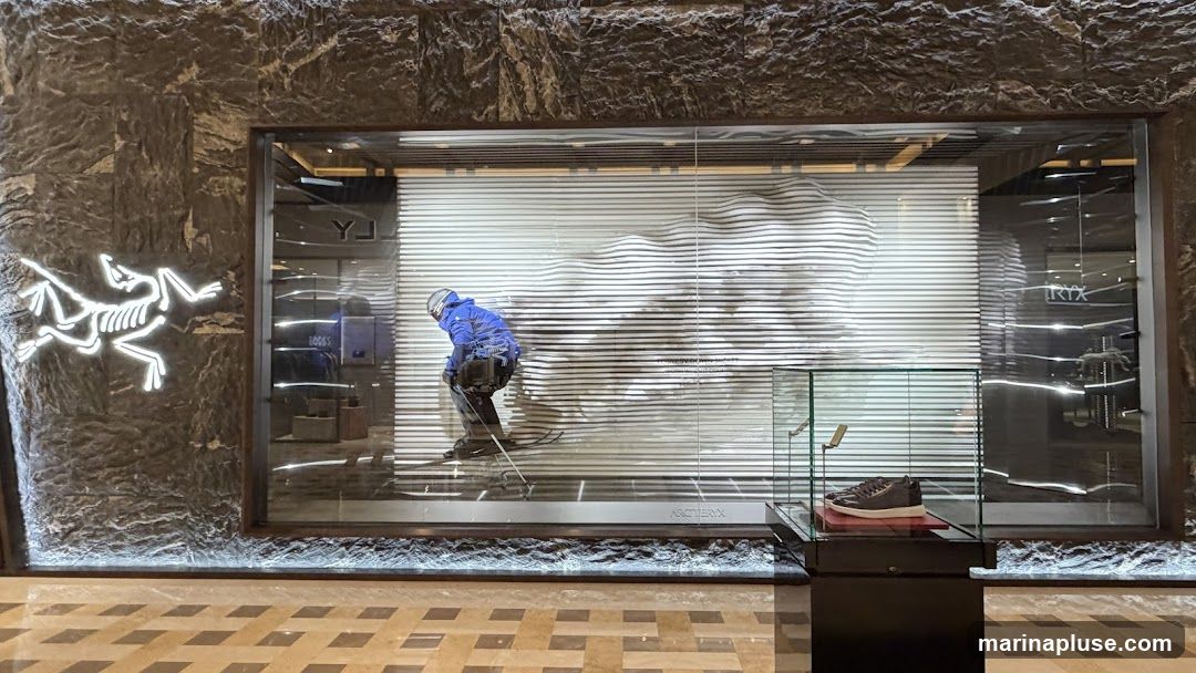 A storefront with a large window display featuring a mannequin dressed in ski gear, appearing to ski down a wavy, striped mountain. A neon Arc'teryx logo is visible on the left side of the stone facade. Inside the window, a glass display case shows a pair of brown shoes. The store name "ARC'TERYX" is visible on a plaque below the display.