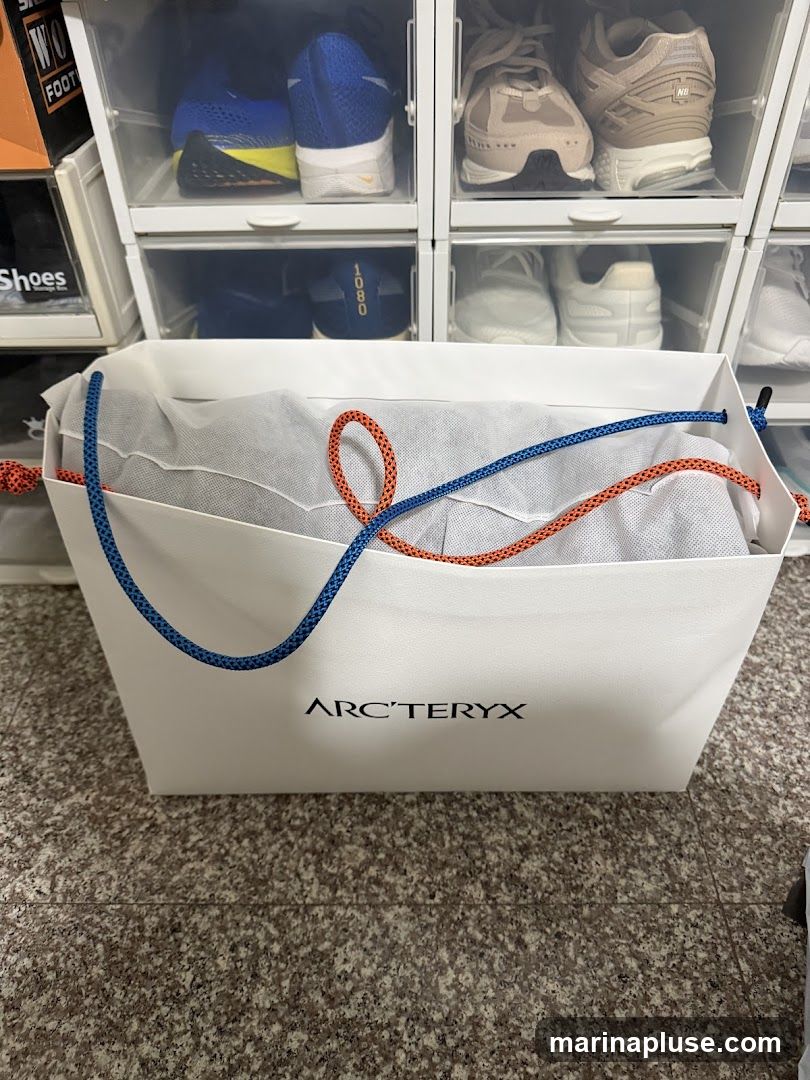A white Arc'teryx shopping bag with blue and orange rope handles is filled with a white garment bag. Behind the shopping bag, several clear shoe storage boxes are stacked, containing various athletic shoes. The floor is a speckled granite pattern.