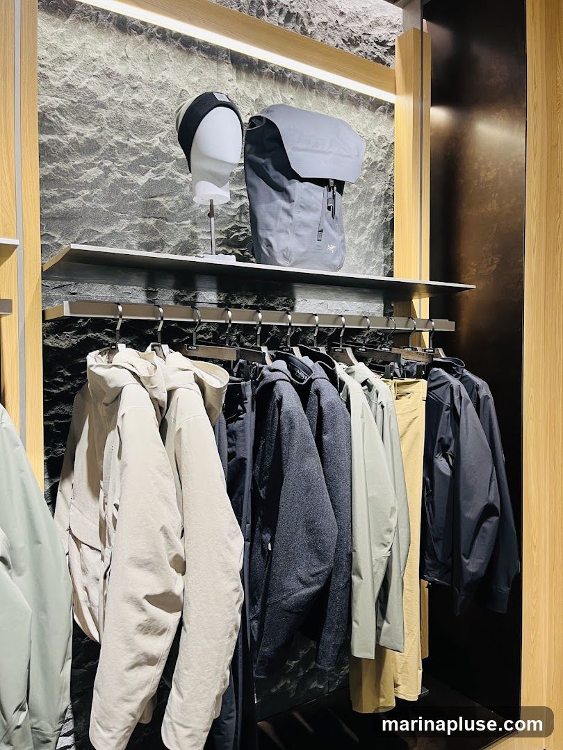 A display of outdoor clothing and accessories, including jackets, pants, a backpack, and a hat on a mannequin head. The clothing is neatly hung on a rack, with a textured stone-like wall as the backdrop.