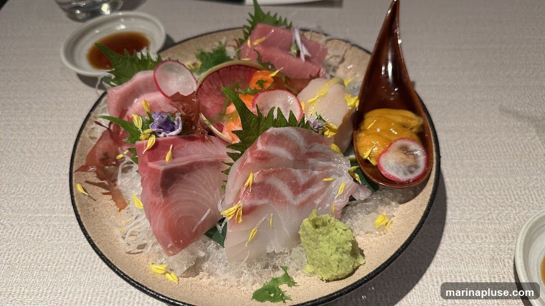 A plate of assorted sashimi, including tuna, hamachi, and uni, garnished with radish, shiso leaves, and edible flowers. A small dish of soy sauce and a mound of wasabi are also visible.