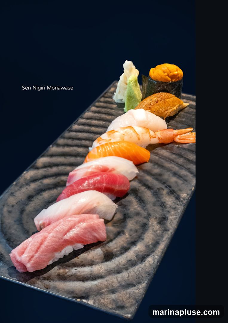 A platter of assorted nigiri sushi is presented on a dark, textured plate against a dark blue background. The sushi includes pieces with tuna, salmon, shrimp, eel, and sea urchin. A small mound of wasabi and pickled ginger is also visible. The text "Sen Nigiri Moriawase" is overlaid on the left side of the image.