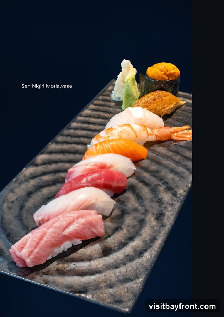 A platter of assorted nigiri sushi is presented on a dark, textured plate against a dark blue background. The sushi includes pieces with tuna, salmon, shrimp, eel, and sea urchin. A small mound of wasabi and pickled ginger is also visible. The text "Sen Nigiri Moriawase" is overlaid on the left side of the image.