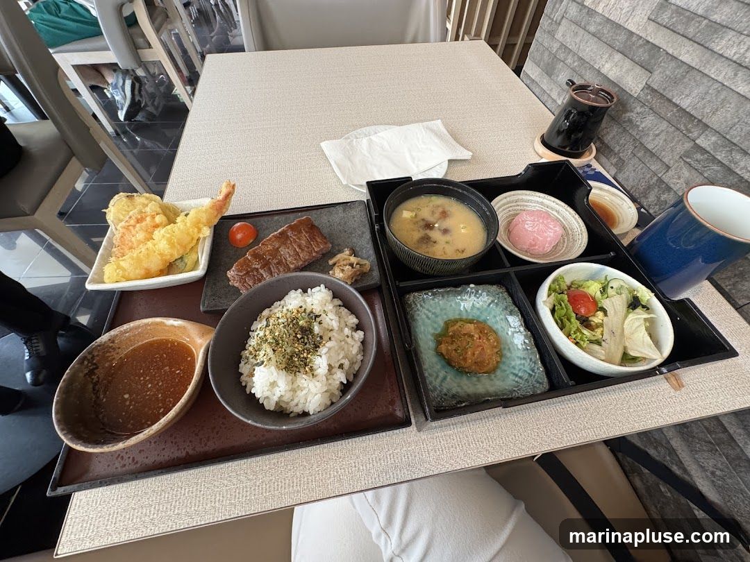 A Japanese meal set is presented on a table. The meal includes tempura, a slice of steak, rice, miso soup, a small salad, and a pink mochi dessert. A dipping sauce and a cup of tea are also provided.