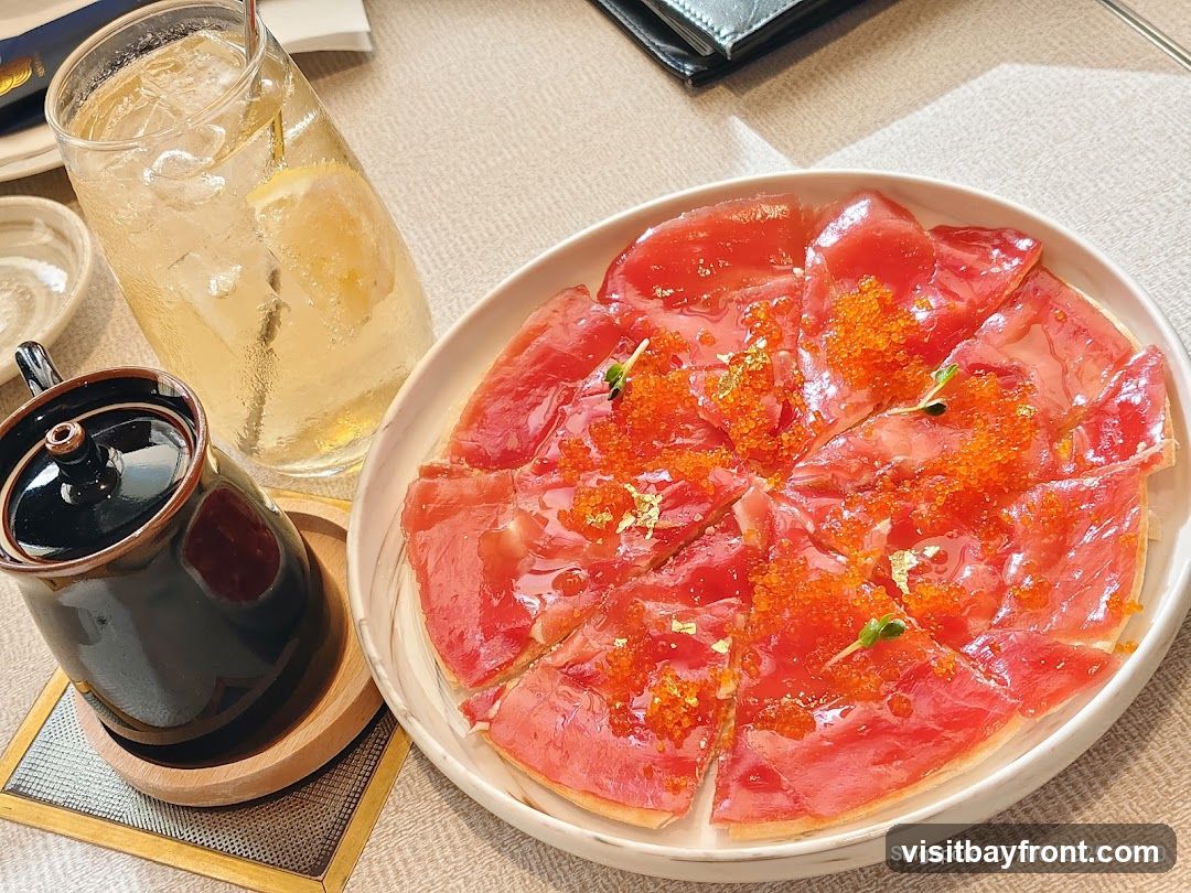 A close-up shot of a tuna tartare pizza, garnished with roe and gold leaf, served on a white plate. A glass of iced beverage with lemon and a small ceramic pot are also visible on the table.