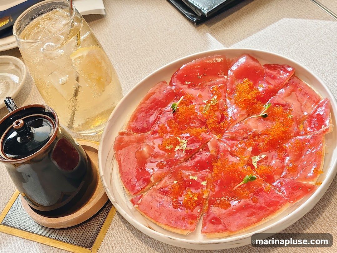 A close-up shot of a tuna tartare pizza, garnished with roe and gold leaf, served on a white plate. A glass of iced beverage with lemon and a small ceramic pot are also visible on the table.