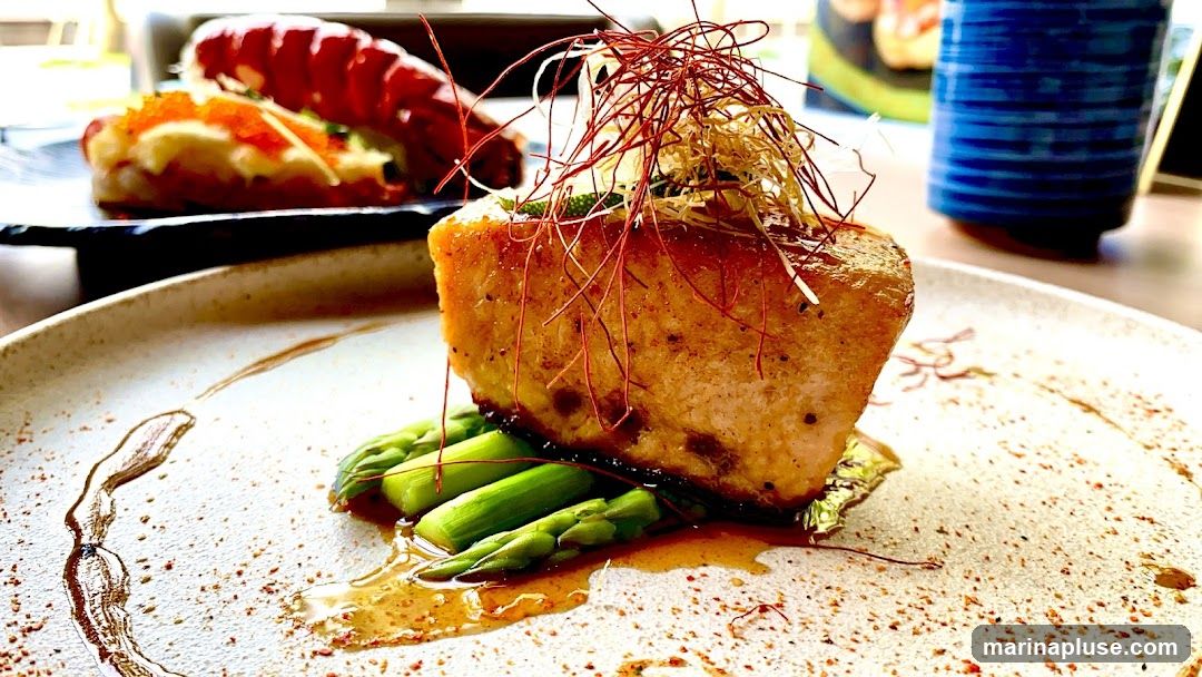 A close-up shot of a piece of seared salmon served with asparagus on a speckled plate. The salmon is topped with shredded red and yellow garnishes. In the background, a lobster tail dish and a blue patterned cup are visible.