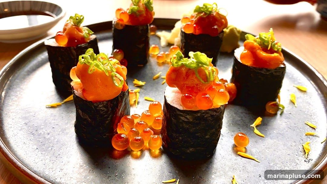 A close-up shot of six pieces of gunkan maki sushi, topped with chopped salmon and ikura (salmon roe), garnished with chopped scallions and served on a dark plate with a small dish of soy sauce in the background.