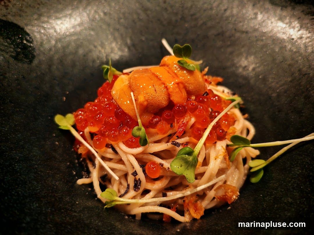 A close-up shot of a bowl of pasta topped with sea urchin and salmon roe. The pasta appears to be thin and light-colored, possibly soba noodles. It is garnished with microgreens and small crispy bits, possibly fried shallots or garlic. The dish is presented in a dark, textured bowl.