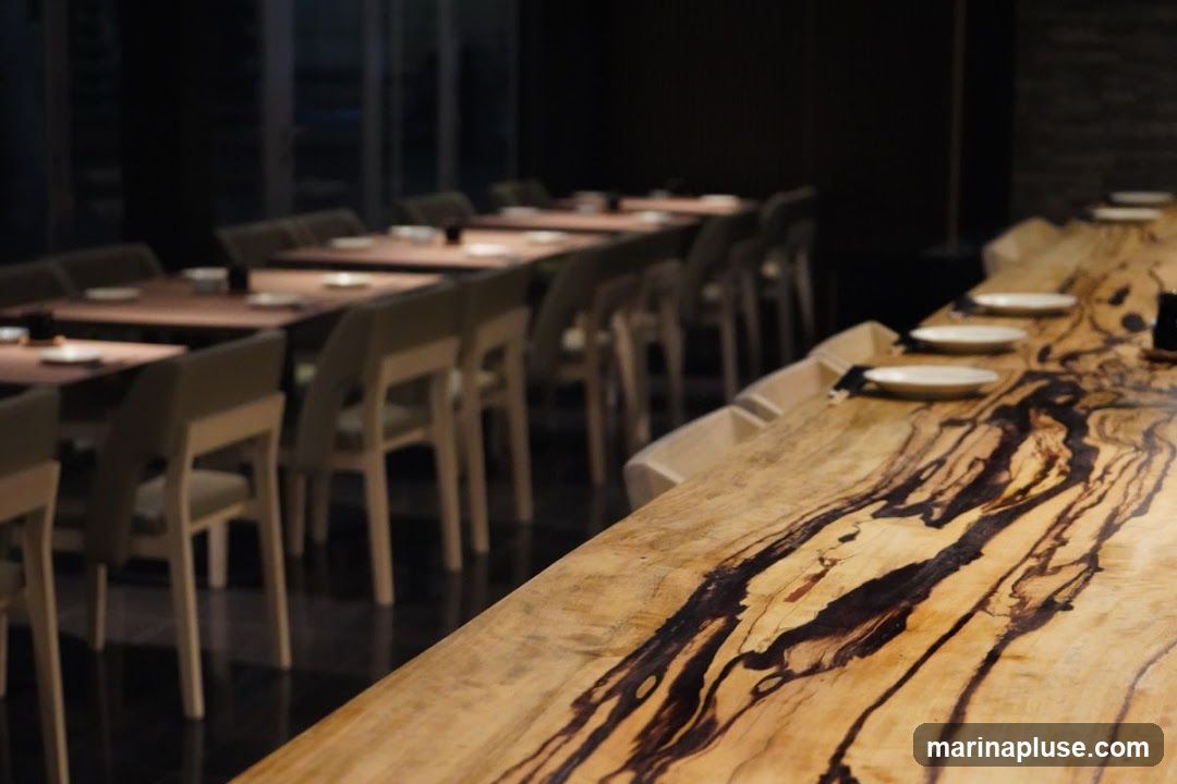 A dimly lit restaurant interior featuring a long, live-edge wooden table in the foreground with plates set. In the background, rows of dining tables and chairs are visible, also set for a meal. The ambiance suggests a sophisticated dining experience.