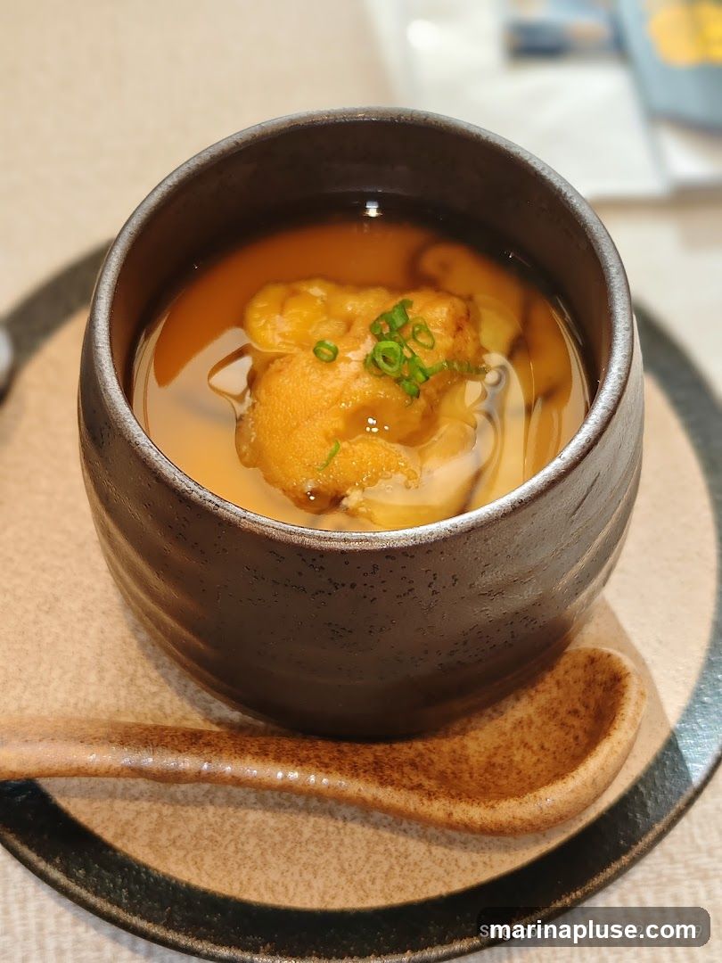 A close-up shot of a bowl of chawanmushi, a Japanese savory egg custard. The custard is topped with uni (sea urchin) and garnished with chopped green onions. A ceramic spoon rests on the side of the bowl, which is placed on a textured plate. The background is blurred, suggesting an indoor dining setting.
