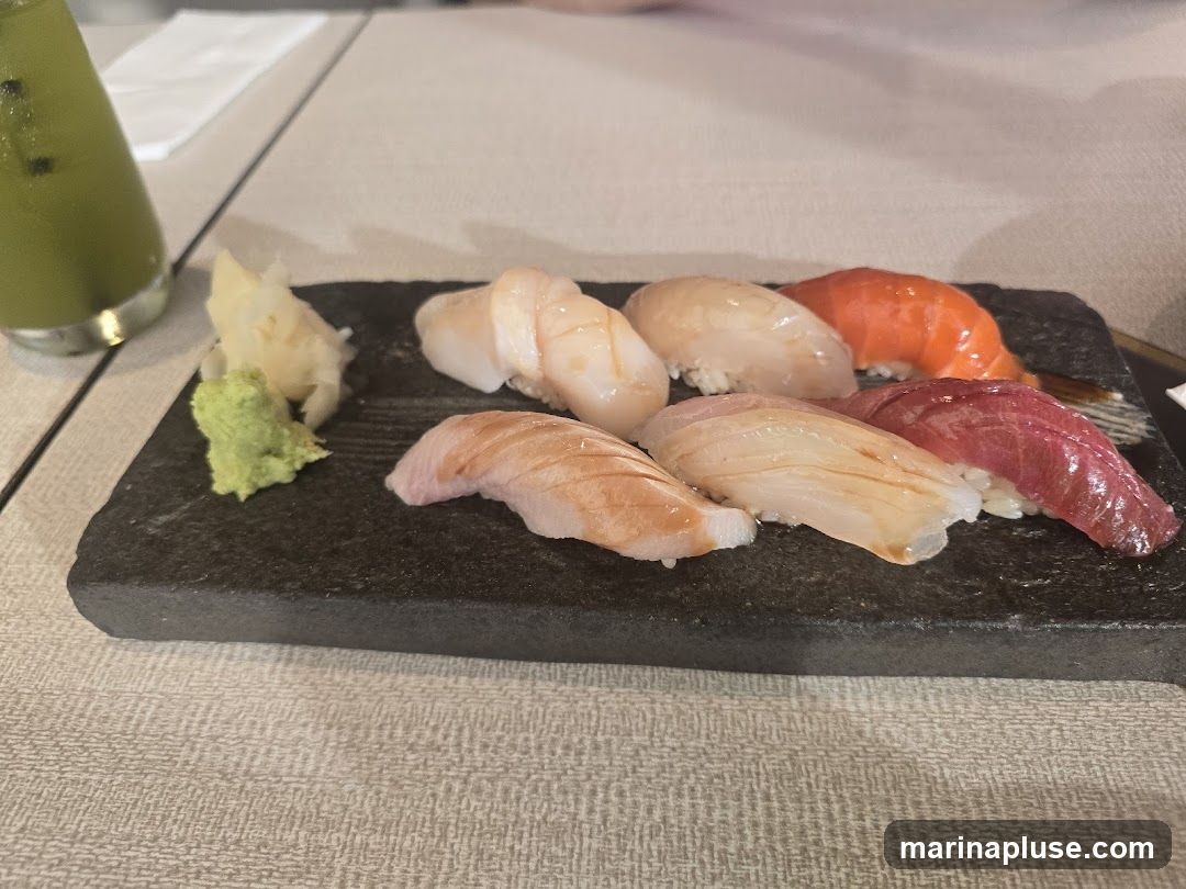 A dark rectangular plate holds a selection of sushi, including salmon, tuna, and white fish. A small mound of wasabi and pickled ginger are also on the plate. A green beverage in a glass is visible to the left.