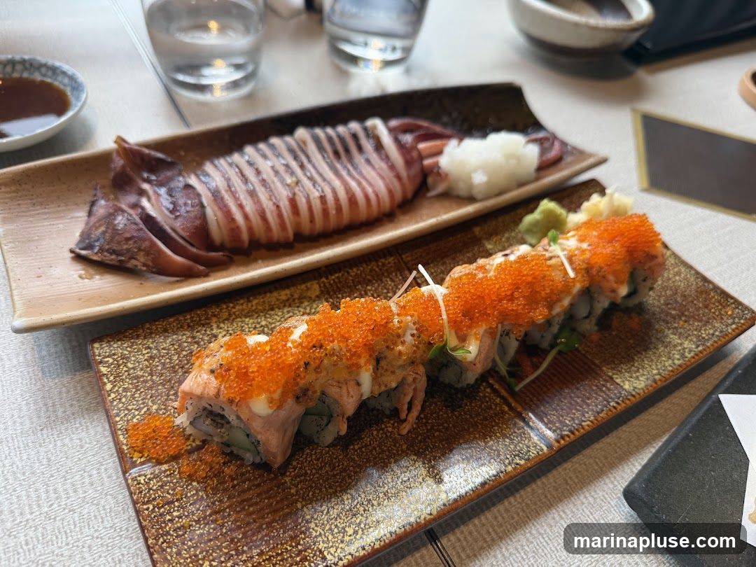 A close-up shot of a meal featuring sushi rolls topped with orange roe and a side of grilled squid. The sushi is presented on a patterned rectangular plate, while the squid is served on a long, light-colored dish with a side of grated daikon radish. A small dish of soy sauce and glasses of water are also visible on the table.