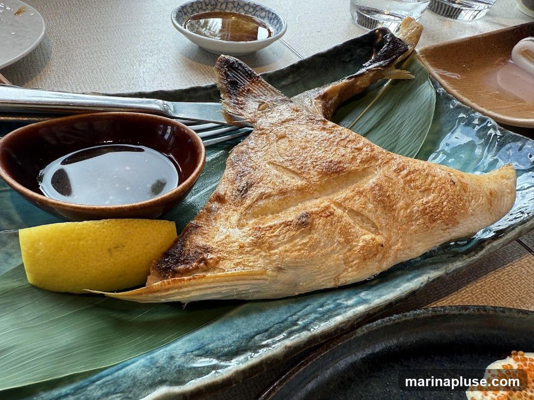 A close-up shot of a grilled fish dish served on a blue-green ceramic plate. The fish is golden brown and appears to be a thick cut, possibly a steak or a large portion of a whole fish. It is garnished with a large green leaf and a wedge of lemon. A small brown bowl contains a dark dipping sauce, and a fork and knife are visible. In the background, there are other dishes and glasses on a table.