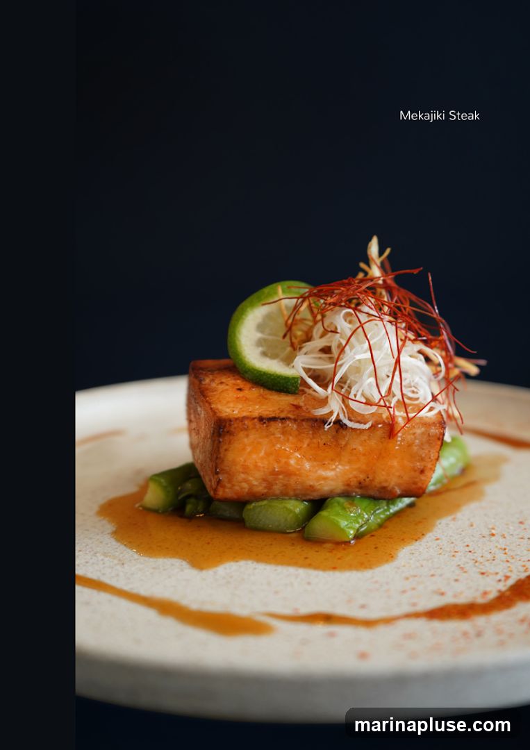 A close-up shot of a perfectly cooked Mekajiki steak served on a bed of asparagus with a drizzle of sauce. The steak is topped with thinly sliced white onions, red chili threads, and a lime wedge.