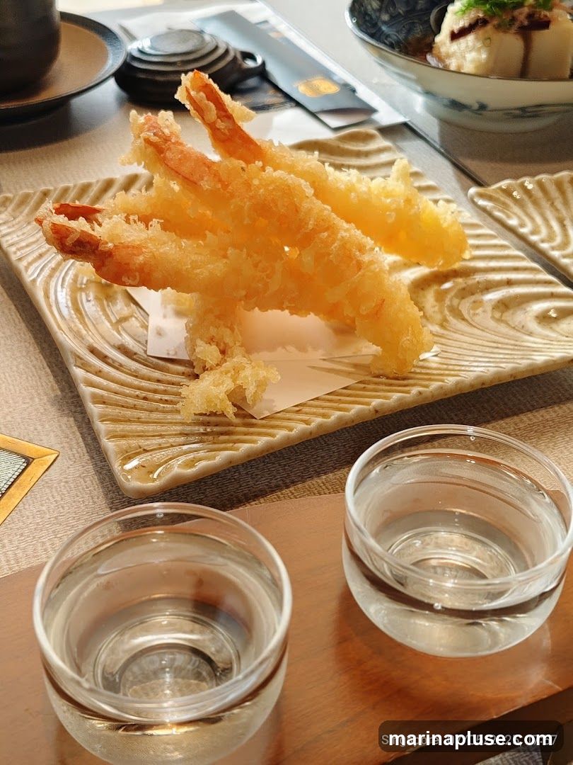 A close-up shot of three pieces of golden-brown shrimp tempura served on a textured, light-colored square plate. The tempura is crispy and appears to be lightly battered. In the foreground, two small, clear glasses filled with a clear liquid, possibly sake or water, are placed on a wooden surface. In the background, a bowl of food with a light-colored topping and a dark sauce is visible, along with a tea set and chopsticks. The overall setting suggests a Japanese dining experience.