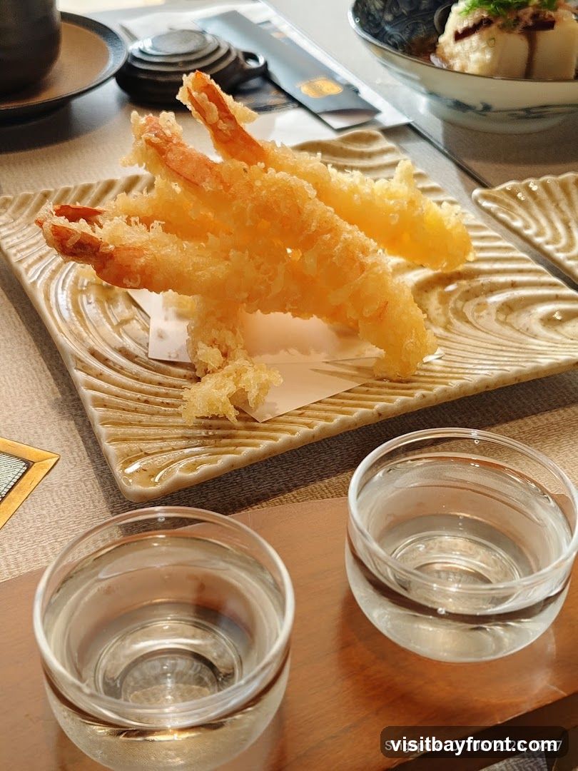 A close-up shot of three pieces of golden-brown shrimp tempura served on a textured, light-colored square plate. The tempura is crispy and appears to be lightly battered. In the foreground, two small, clear glasses filled with a clear liquid, possibly sake or water, are placed on a wooden surface. In the background, a bowl of food with a light-colored topping and a dark sauce is visible, along with a tea set and chopsticks. The overall setting suggests a Japanese dining experience.