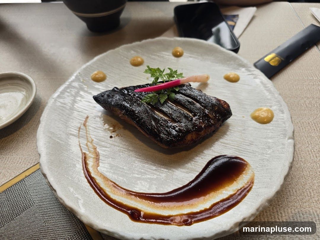 A close-up shot of a grilled fish dish served on a textured white plate. The fish is dark and appears to be glazed, with visible grill marks. It is garnished with a sprig of parsley and a pink pickled element. The plate is decorated with dollops of yellow sauce and a swirl of dark sauce, creating a smiley face design. A small dish and a black chopstick holder are visible in the background.
