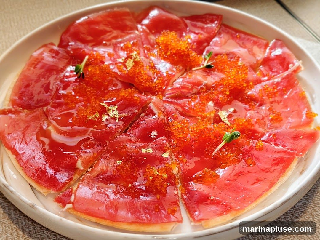 A close-up shot of a circular dish filled with thinly sliced raw tuna, topped with bright orange roe, a glistening sauce, and delicate gold leaf flakes. Small green sprouts are used as garnish. The dish is presented on a white plate with a textured rim, set against a neutral background.
