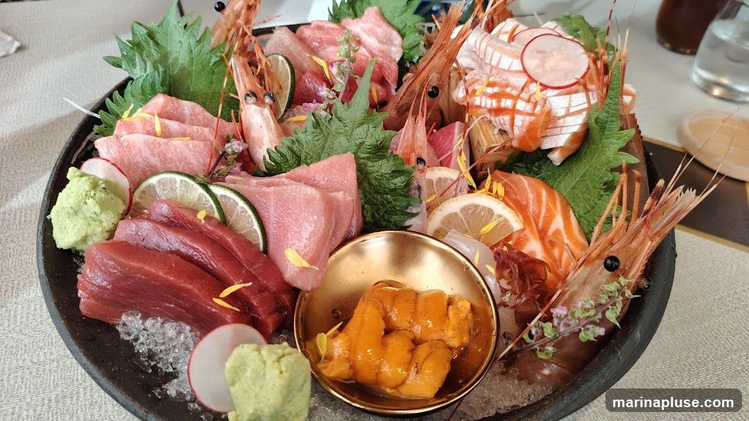 A large platter of assorted sashimi, including tuna, salmon, and shrimp, served with wasabi, lime, and uni (sea urchin). The sashimi is arranged on a bed of crushed ice and garnished with shiso leaves and edible flowers.