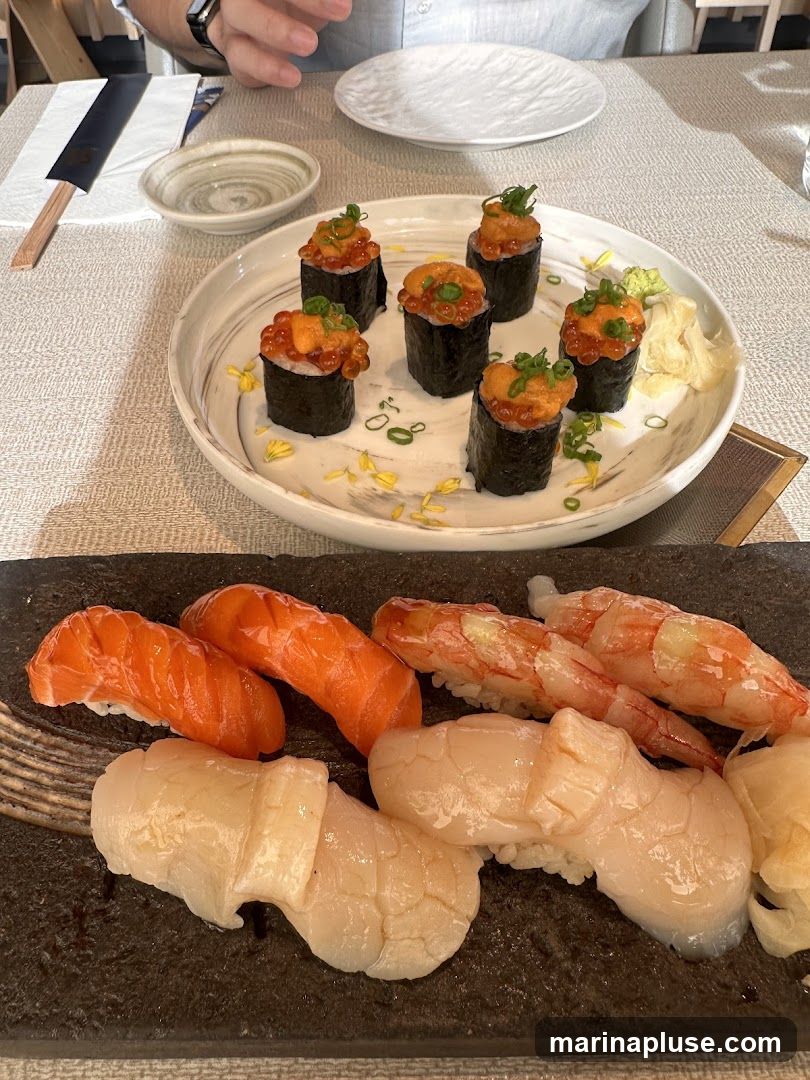 A close-up shot of two platters of sushi. The top platter contains six pieces of gunkan maki sushi, each topped with ikura (salmon roe) and uni (sea urchin), garnished with chopped scallions and yellow flower petals. The bottom platter features several pieces of nigiri sushi, including salmon, shrimp, and scallop, served with pickled ginger.