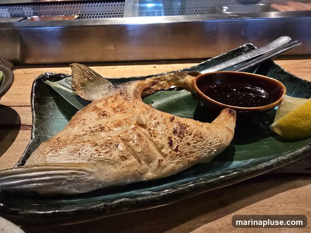 A close-up shot of a grilled fish dish served on a dark green, textured plate. The fish appears to be a large portion, possibly a steak or half of a fish, with visible grill marks and crispy skin. It is garnished with a lemon wedge and a small bowl of dark dipping sauce. A banana leaf is placed under the fish. In the background, a stainless steel counter and wooden surfaces suggest a restaurant setting.