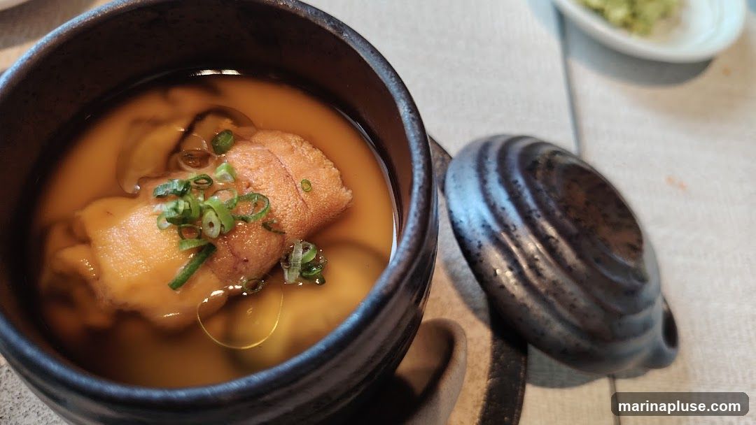 A close-up shot of a Japanese savory egg custard dish called Chawanmushi, topped with uni (sea urchin) and chopped scallions. The dish is served in a dark ceramic bowl with a lid. A small dish of green paste, possibly wasabi or a condiment, is visible in the background.