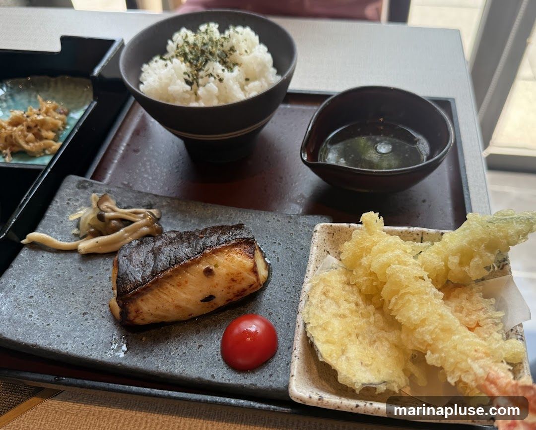 A Japanese meal served on a tray. It includes a bowl of white rice topped with green flakes, a small dish of sauce, a piece of grilled fish with mushrooms and a cherry tomato, and a side of tempura with shrimp and vegetables. Another small dish with a side dish is visible on the left.