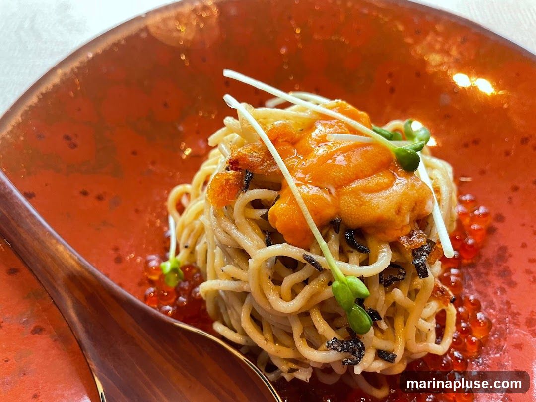 A close-up shot of a noodle dish served in a rustic orange bowl. The noodles are topped with bright orange sea urchin (uni) and garnished with microgreens and what appears to be crispy fried shallots or garlic. Red ikura (salmon roe) is scattered around the base of the noodles. A wooden spoon is visible on the left side of the bowl.