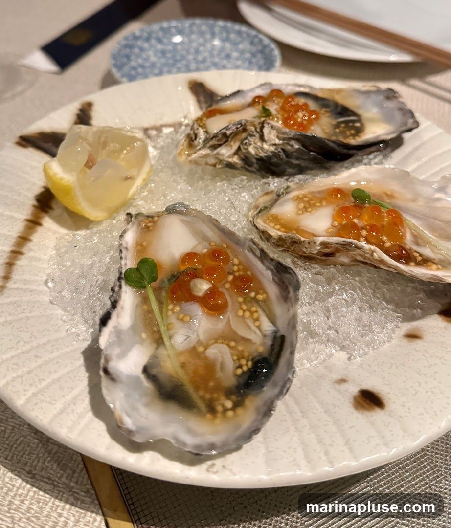 A plate of three oysters served on crushed ice. Each oyster is topped with roe, small seeds, and a garnish. A wedge of lemon is placed on the side of the plate.
