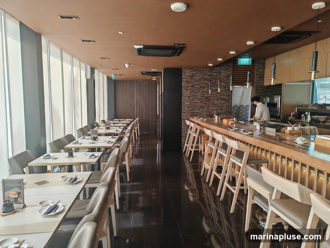 The interior of a modern Japanese restaurant with a sushi bar and dining tables. The dining area features long tables set with place settings and a view of the outdoors through large windows with blinds. The sushi bar is made of light-colored wood with stools for seating. A chef is visible behind the bar preparing food. The overall ambiance is clean and contemporary.