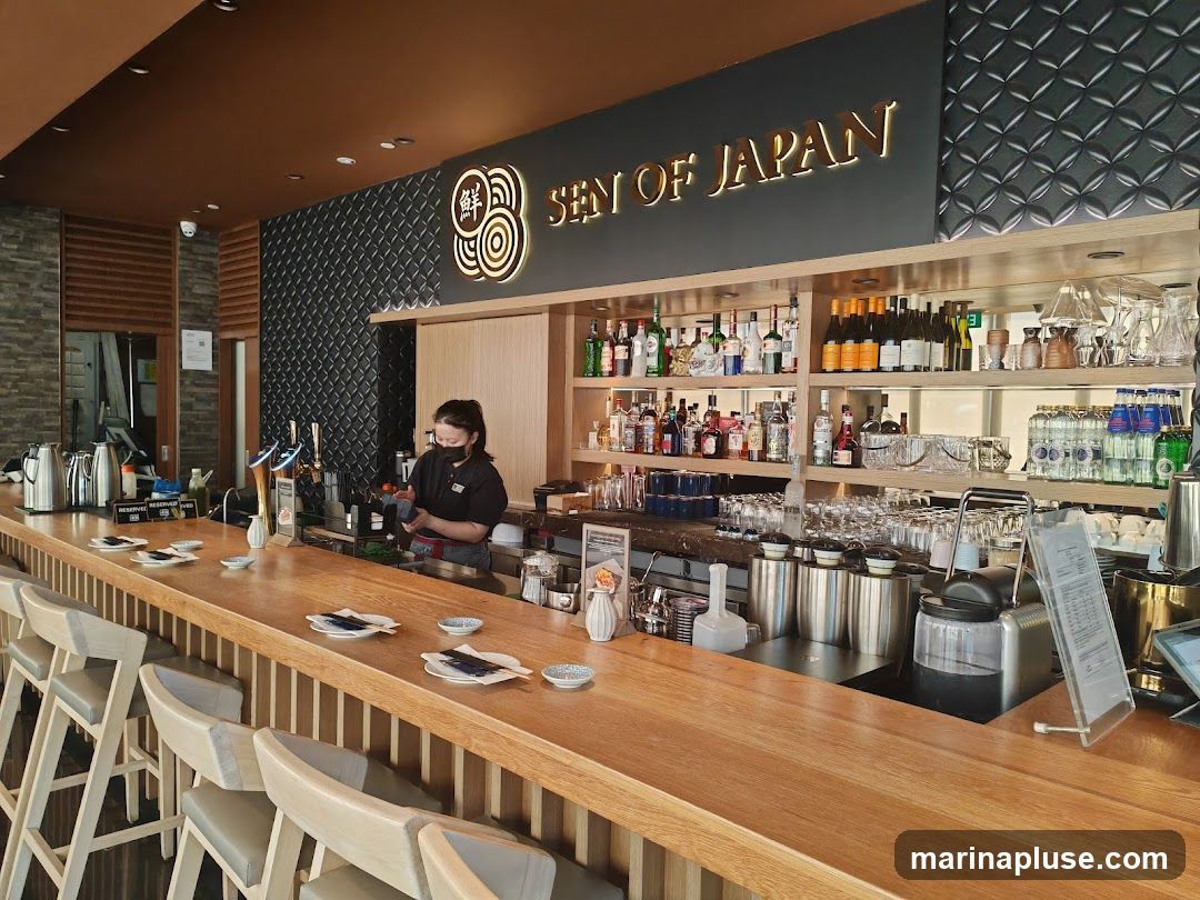 The image shows the interior of a bar or restaurant with a long wooden counter. A bartender wearing a mask is visible behind the counter, preparing drinks. Shelves behind the bar are stocked with various bottles of alcohol and glassware. The establishment's name, "SEN OF JAPAN," is displayed prominently on the wall above the bar in illuminated golden lettering. Several bar stools are arranged along the counter.