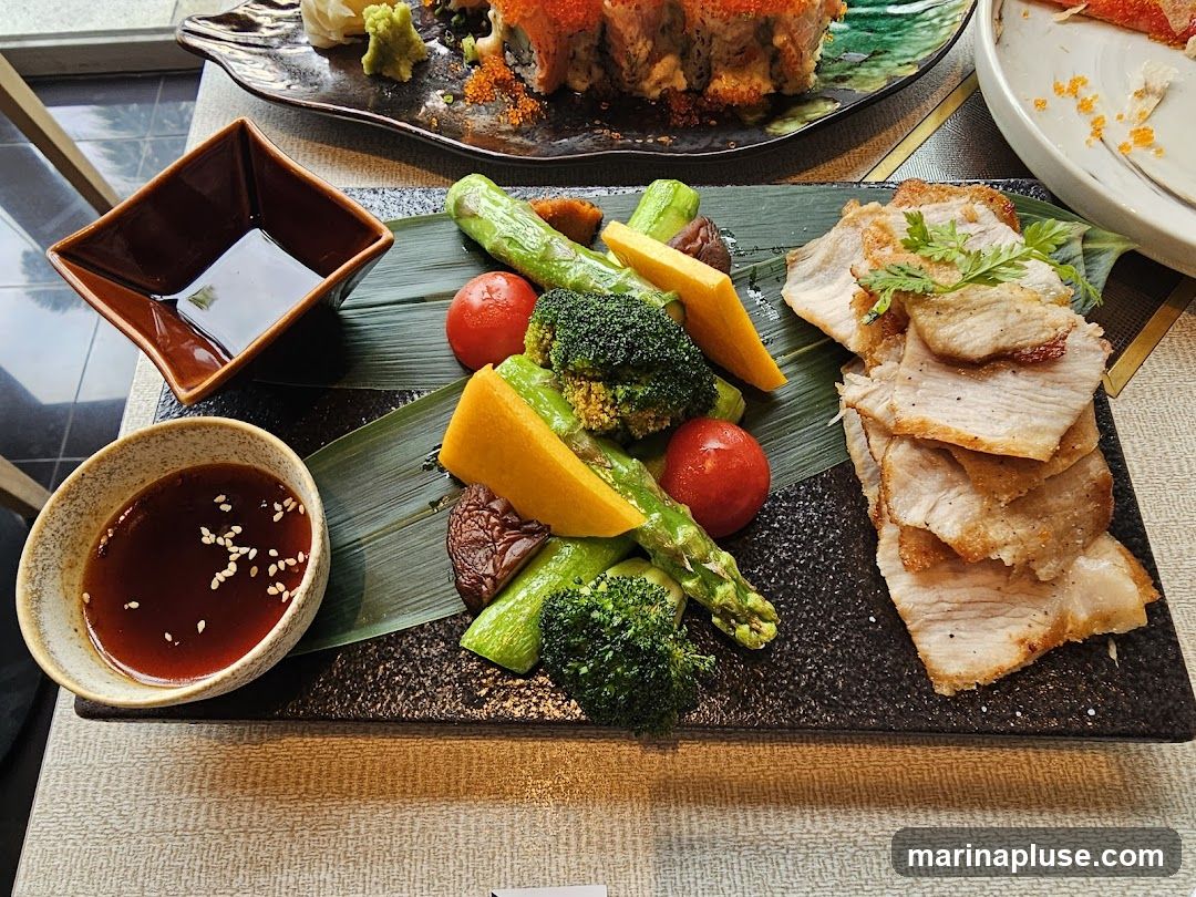 A platter of thinly sliced, pan-fried pork belly is served alongside a colorful assortment of steamed vegetables, including broccoli florets, asparagus spears, cherry tomatoes, and chunks of yellow squash. Two dipping sauces are provided: one in a small, speckled bowl with sesame seeds, and another in a dark, square dish. In the background, a plate of sushi rolls is visible.