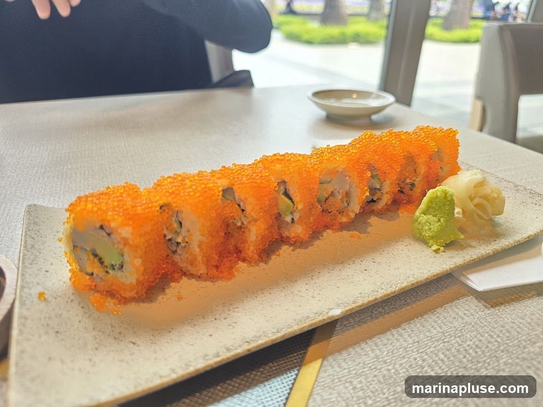 A plate of California rolls covered in orange tobiko, served with wasabi and pickled ginger. The rolls are arranged in a line on a rectangular plate. In the background, a person's hands are visible, and a small dish and a blurred outdoor scene can be seen.