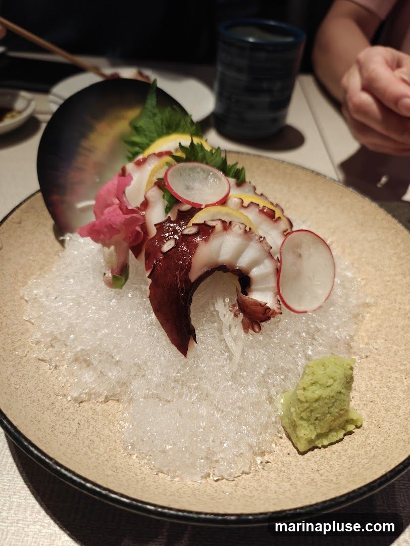 A plate of octopus sashimi served on crushed ice, garnished with lemon slices, radish, and a dollop of wasabi. A dark decorative element is behind the sashimi. In the background, a person's hands are visible, along with a dark blue cup and chopsticks.