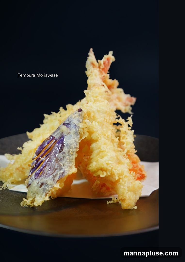 A close-up shot of a plate of tempura, featuring shrimp and eggplant. The food is golden brown and crispy, presented on a dark plate with a white paper liner. The text "Tempura Moriawase" is overlaid on the dark background.