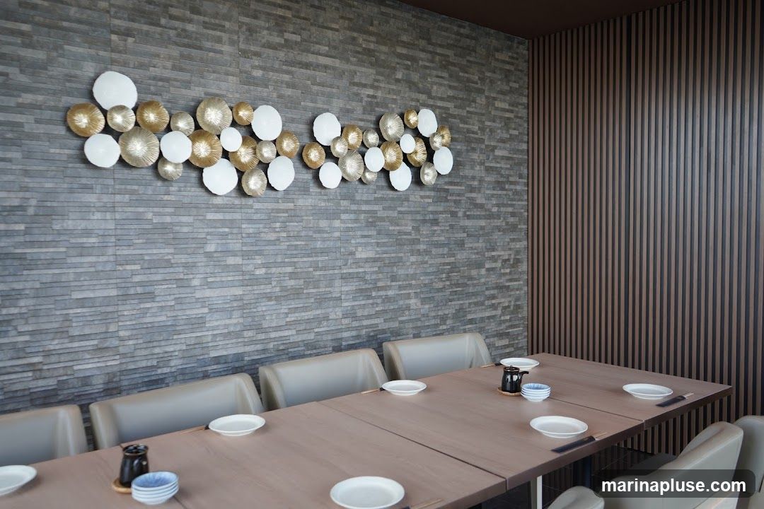 A long wooden dining table is set with plates, bowls, and chopsticks. The table is in a room with a stone accent wall featuring a decorative metal art piece and a wall with vertical wooden slats.