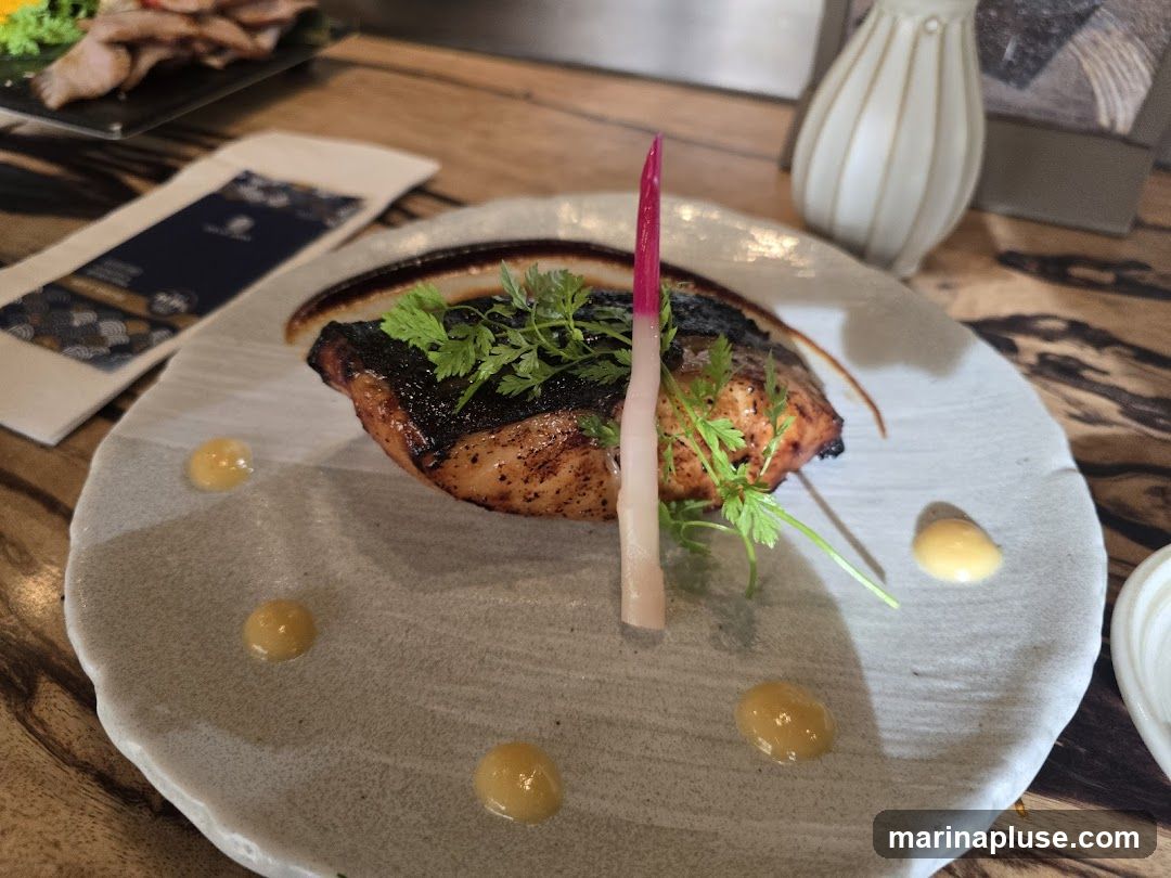 A close-up shot of a beautifully plated fish dish. The fish, with a dark, crispy exterior, is garnished with fresh green herbs and a vibrant pink and white pickled vegetable. It is served on a textured, light-colored plate with dollops of a yellow sauce. In the background, a blurred menu or placemat with a dark blue and gold pattern is visible, along with a portion of another dish and a decorative vase. The table surface appears to be made of wood.