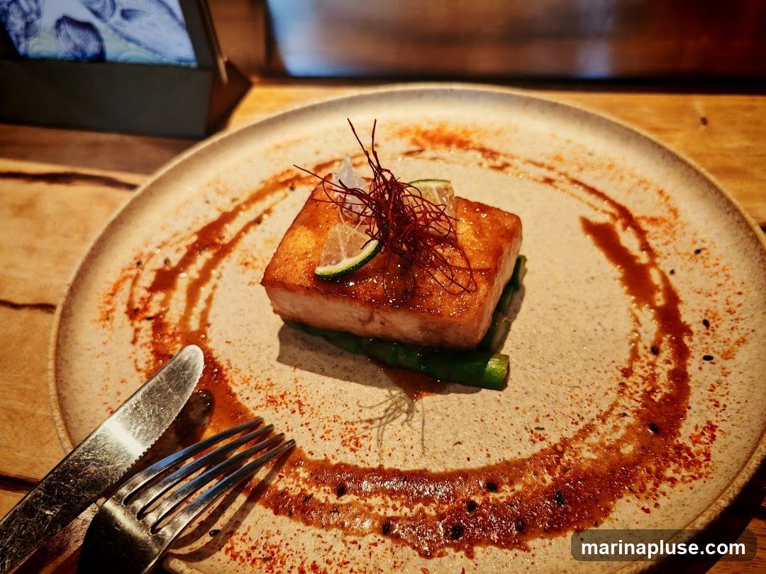 A close-up shot of a beautifully plated salmon dish. The salmon fillet is glazed and sits atop a bed of green asparagus. It is garnished with lime slices and thin red threads. The plate is decorated with a swirl of sauce and sprinkled with spices. A fork and knife are placed on the side of the plate. The background shows a wooden table and a blurred image of what appears to be a menu or artwork in a stand.
