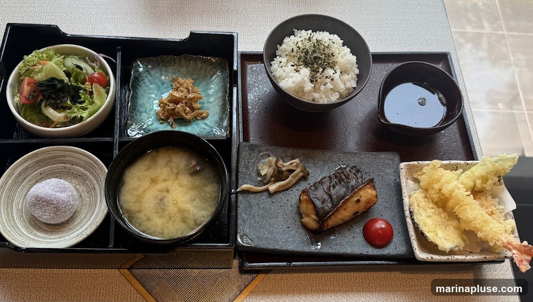 A Japanese set meal is presented in a black bento box. The meal includes a bowl of white rice topped with furikake, a small dish of soy sauce, a piece of grilled fish, a cherry tomato, mushrooms, tempura (shrimp and eggplant), a bowl of miso soup, a small dish of pickled vegetables, a salad with lettuce and tomatoes, and a round dessert dusted with powder.