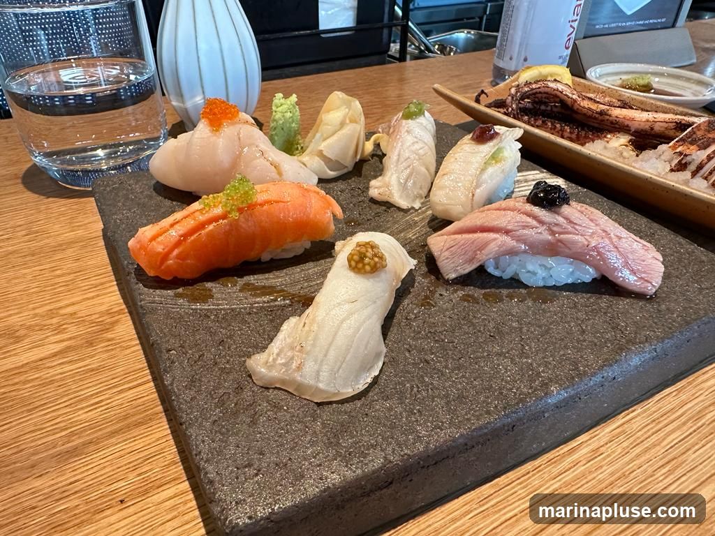 A close-up shot of a variety of sushi pieces arranged on a dark stone platter. The sushi includes salmon with roe, scallop with roe, white fish with a dollop of sauce, and a piece of seared tuna with black caviar. In the background, a glass of water, a decorative vase, and a dish with grilled octopus and rice are visible. The setting appears to be a restaurant table.
