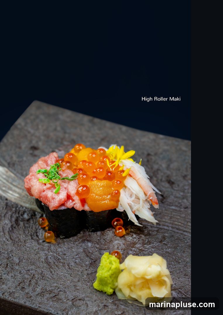 A close-up shot of a sushi roll topped with minced tuna, salmon roe, uni, and crab meat. It is served on a dark, textured plate with a side of wasabi and pickled ginger. The text "High Roller Maki" is visible in the upper right corner.