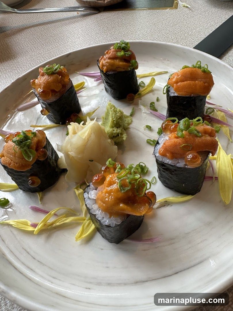 A plate of six pieces of uni gunkan maki sushi, garnished with ikura (salmon roe) and chopped scallions. The sushi is served with a side of pickled ginger and wasabi on a marbled plate, decorated with flower petals.