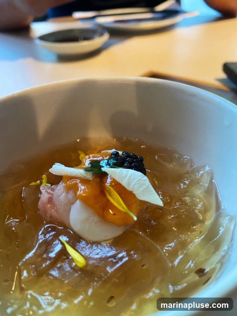 A close-up shot of a delicate appetizer served in a white bowl. The dish features a clear, gelatinous base with pieces of seafood, topped with a dollop of orange sauce, black caviar, and a white garnish, possibly a flower petal. Yellow flower petals are scattered on top and within the jelly. The background shows a blurred dining setting with plates and chopsticks.